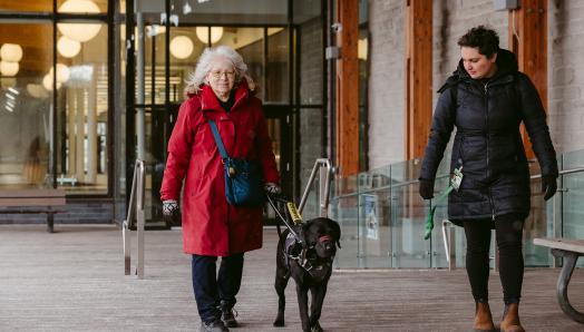 Cheri sort d’un immeuble vitré, guidée par Sassy, son chien-guide d’INCA, une chienne labrador retriever noire qui marche à sa gauche. L’intervenante de Cheri marche à gauche de la chienne en l’obsevant. Les femmes portent des manteaux d’hiver.