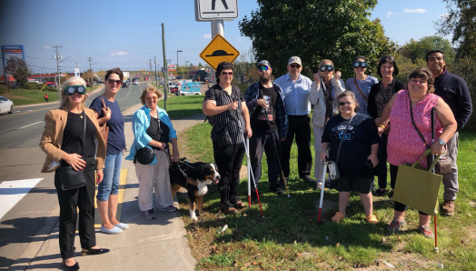 Un groupe de personnes ayant une limitation visuelle se tient à côté d'un passage pour piétons avec des agents publics, à Moncton lors de la campagne « Montez à bord » de l'automne 2023.