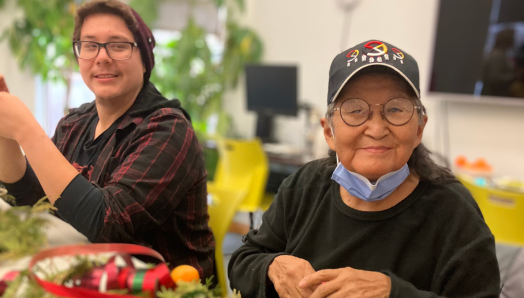 Deux participants autochtones souriants sont assis à une table au bureau d'INCA à Thunder Bay. Au cours d'un programme de vacances, elles créent des décorations avec des nœuds de cèdre frais, des tranches d'orange séchées, des biscuits de pain d'épice faits maison, des rubans, des cloches, des bâtons de cannelle et de la ficelle de jute.