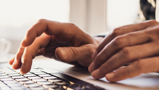Gros plan sur les mains d'une personne tapant sur le clavier d'un ordinateur portable. L'accent est mis sur les mains et le clavier, l'arrière-plan est flou mais il s'agit d'un intérieur bien éclairé
