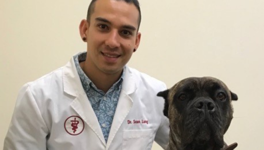 Dr Lang s’agenouille sur le sol à côté de son chien, Adelina. Dr Lang porte une blouse blanche de vétérinaire. Adelina est une chienne de taille moyenne, de couleur noire et marron foncé.