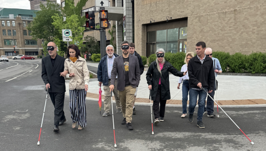 Claude les yeux bandés guidé par Valérie directrice d’INCA Québec, Pierre-Luc les yeux bandé guidé par Jean-Michel et un bénévole derrière lui, Maude guidée par David et René du RPHV 03-12 à l’arrière traversent la rue en direction de la caméra en balayant leur canne-blanche devant eux