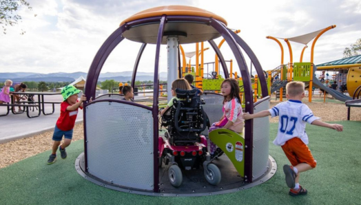 Des enfants jouent sur une aire de jeux accessible par une journée d'été ensoleillée. Un enfant en fauteuil roulant se trouve sur un carrousel accessible aux fauteuils roulants avec trois autres enfants. Deux autres enfants courent et tirent le carrousel pour le faire tourner pour les enfants assis dessus.