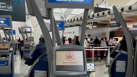 Un kiosque d'enregistrement libre-service à l'aéroport Pearson. L'écran du kiosque affiche le logo d'Air Canada et, au-dessus, un panneau indique : « Mode basse vision disponible sur ce kiosque ». Un panneau avec des boutons de navigation accessibles est situé sur le côté droit de la borne, à côté de l'écran tactile.
