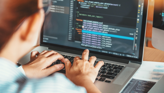 Une personne tape sur un ordinateur portable. La photo est prise par-dessus son épaule et l'écran de son ordinateur portable affiche des lignes de code, ce qui suggère qu'elle est en train de programmer ou de coder.
