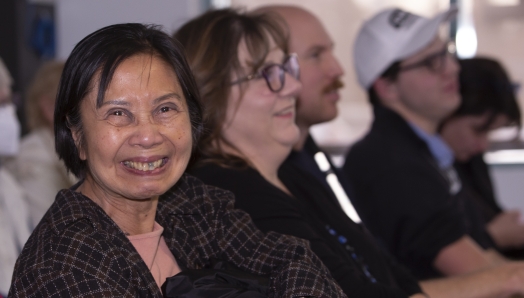 Une femme ayant une perte de vision regarde directement la caméra et sourit. Elle est assise au milieu d'un grand groupe de personnes hors champ en arrière-plan