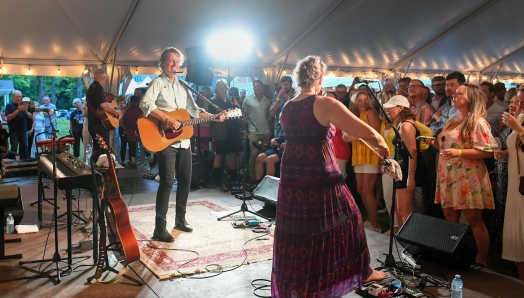 Guitare à la main, Jim Cuddy se tient sur une scène. Derrière lui, un autre homme joue de la guitare. À sa gauche, une femme portant une robe mauve joue du violon. Une foule dense se tient devant eux.
