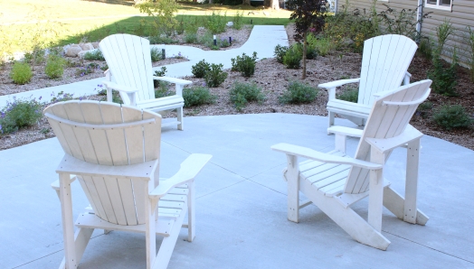 Quatre chaises de Muskoka blanches sont disposées en cercle sur une surface en ciment. Un sentier traverse des parterres de jardin avec de petits arbres et arbustes. 