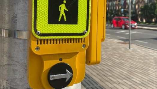 Une signalisation piétonnière accessible (SPA) est montée sur un poteau métallique à un passage pour piétons à Ottawa. La zone environnante montre un environnement urbain avec un trottoir pavé de briques, une voiture rouge en arrière-plan et des immeubles modernes sous un ciel bleu clair.