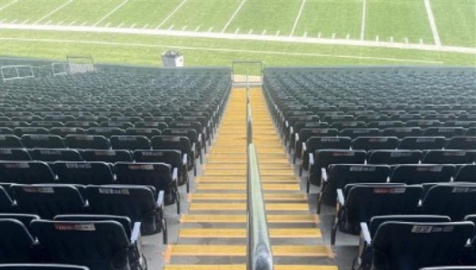 Une volée d’escaliers de stade en béton, dont le sommet donne sur un terrain de football, avec des rangées de sièges en gradins de part et d’autre. Les escaliers sont séparés au milieu par une rampe et chaque escalier est peint d’un jaune doré brillant sur toute la longueur des bords avant.