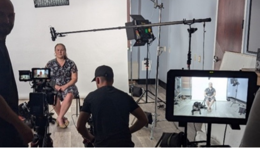 Taylor Bauer et son chien-guide Wallace sont interviewés aux fins d’une vidéo d’éducation publique. Taylor est assise avec un écran blanc derrière elle, tandis que plusieurs membres du personnel de production font fonctionner une caméra et du matériel d’enregistrement devant elle.