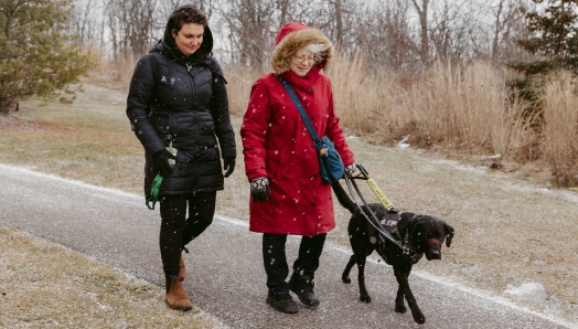 Cheri, qui porte une parka rouge à capuche, marche sur un chemin pavé avec Sassy, sa chienne-guide Labrador noire d'INCA, et une intervenante des Services communautaires Surdicécité d'INCA.