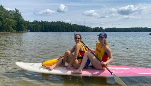 Deux jeunes femmes portant des maillots de surveillantes de baignade sont assises sur une planche à pagaie sur le lac.