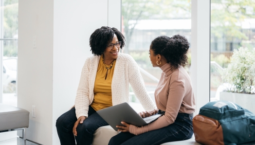 Une femme noire est assise sur un banc et sourit pendant une conversation avec une autre jeune femme noire. Toutes deux sont habillées de manière professionnelle et engagées dans une conversation