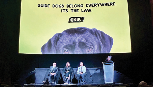 Trois personnes sont assises sur une scène en attente de participer à une table ronde. Un modérateur se tient à leurs côtés, derrière un podium. Une photo de la tête d’un chien-guide avec les mots « Guide dogs belong everywhere. It’s the law » (Les chiens-guides sont à leur place partout. C’est la loi) est projetée derrière eux.