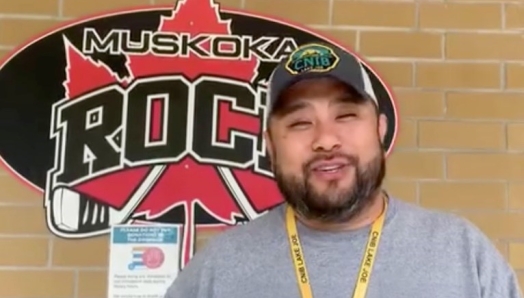 Eugène, qui porte une casquette de baseball, un chandail et un cordon de l'INCA Lake Joe, se tient à l'extérieur d'un bâtiment derrière lequel se trouve le logo " Muskoka Rocks " (un cercle noir avec une feuille d'érable rouge et deux bâtons de hockey).