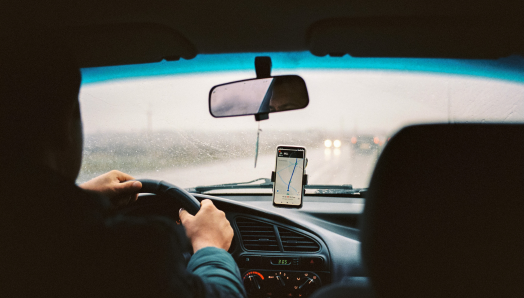 À l'intérieur d'une voiture de covoiturage, la photo est prise du point de vue du passager sur la banquette arrière. Il y a un téléphone intelligent monté sur le tableau de bord de la voiture affichant une application de rideshare. Le conducteur a les mains sur le volant et il pleut à l'extérieur.