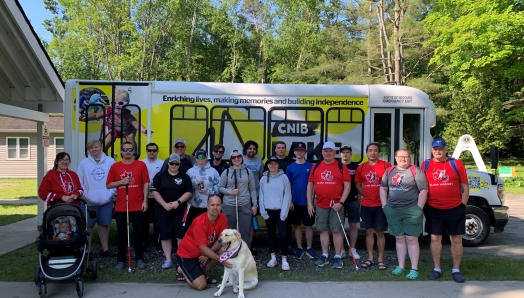 Environ 20 personnes se tiennent devant le nouveau minibus du Camp Lake Joe d'INCA. Plusieurs portent des t-shirts rouges du Hockey sonore Canada et tiennent des cannes blanches. Un chien-guide se trouve devant.