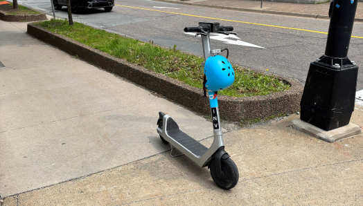 Une trottinette électrique grise est garée au milieu d'un trottoir pour piétons, avec un casque bleu posé à l'avant.