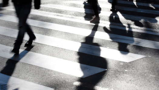 Un passage piéton avec les ombres de personnes qui marchent. L'image est en noir et blanc et montre les jambes de plusieurs personnes qui traversent une rue. 