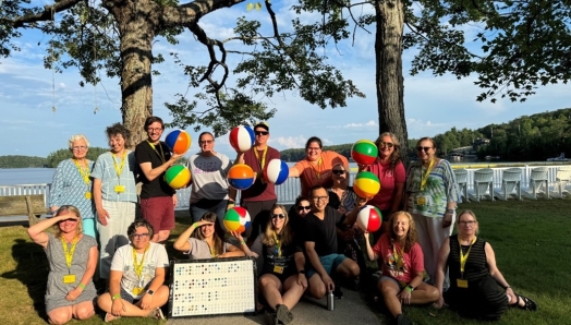 Un groupe d'employés et de bénévoles du Camp Lake Joe d’INCA souriant à l'extérieur par une journée ensoleillée au bord du lac Joseph. Ils tiennent des ballons de plage colorés sur lesquels est imprimé « BRL » pour représenter le mot braille. Ils posent sous de grands arbres avec des chaises blanches et de l'eau en arrière-plan.