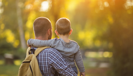 Un père tient son jeune fils dans ses bras. La photo est prise de dos, et l'enfant a son bras autour des épaules de son père. L'arrière-plan est une scène extérieure floue aux tons jaunes et verts.