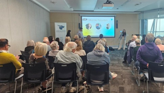 Un groupe de personnes est assis en rangées dans une grande salle de réunion. Vues de dos, elles font face à deux présentateurs à l'avant de la salle, debout à côté d'un écran affichant des diapositives de présentation.