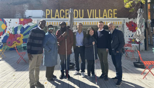 Shalom Foly, Thama Cherilus, Eitel Houédakor, Jean-Pierre Khouzam, Najla Noori, Chris McCray et Cosimo La Rosa posent devant la Place du Village, sous le soleil. Eitel et Najla tiennent leur canne blanche.