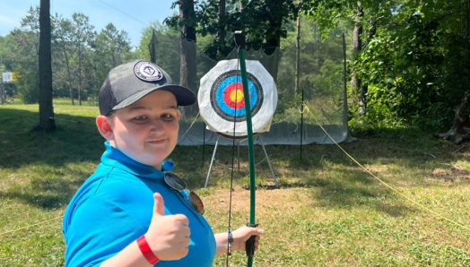 Un garçon tenant un arc et une flèche se tient devant une cible de tir à l'arc, levant le pouce en signe de victoire.