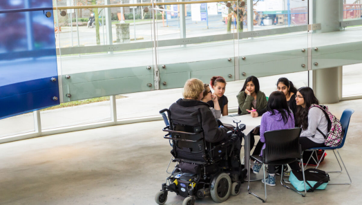 Un groupe de sept jeunes étudiants sont assis ensemble à une table dans une salle de classe et participent à une activité d'apprentissage partagé. L'un des étudiants utilise un fauteuil roulant.