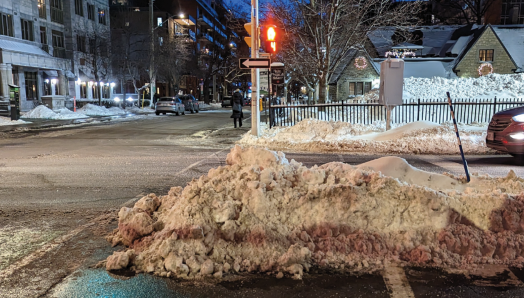 Un passage piéton dans un quartier urbain est montré un soir d'hiver. Le passage piéton est bloqué par un banc de neige de plusieurs mètres de haut.