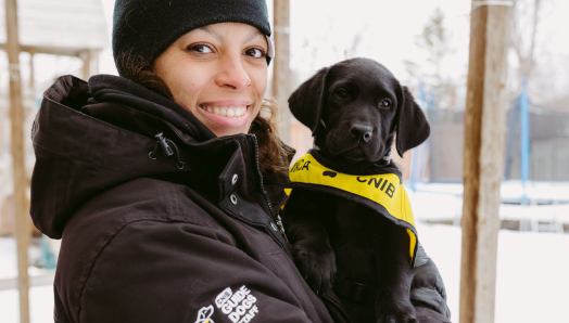 Une personne souriante, vêtue d'une veste d'hiver, tient dans ses bras un chiot noir portant un gilet du programme « Future Guide Dog » d'INCA.