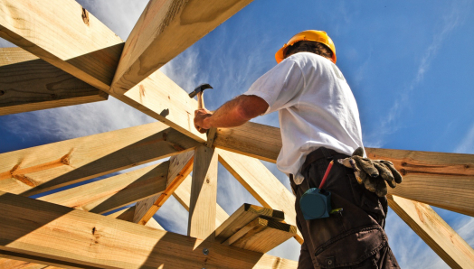 un ouvrier du bâtiment portant un casque de chantier utilise un marteau pour fixer des poutres de toit en bois, vu d'en bas sous un ciel bleu éclatant.
