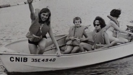 Une photo en noir et blanc montrant deux femmes et deux enfants portant des gilets de sauvetage. Ils sourient, assis dans un bateau sur lequel figure l'inscription « INCA », sur un lac.