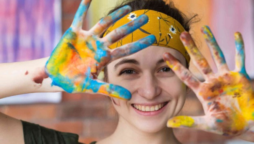 Une jeune femme sourit à la caméra. Ses cheveux sont retenus par un bandana doré à fleurs. Elle tient ses mains ouvertes devant elle, et celles-ci sont recouvertes de peinture aux couleurs vives.