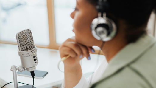 Une femme portant un casque audio est assise à un bureau, à côté d'un micro sur pied. Elle est en train d'enregistrer un podcast.