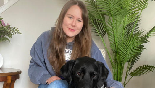 Taylor, une jeune femme aux longs cheveux bruns, est assise sur une chaise, le bras autour de son chien-guide Wallace, un labrador noir portant un harnais d'INCA, qui est assis à ses côtés.