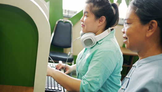Dans un bureau cloisonné, une femme est assise devant un poste de travail informatique et tape sur un clavier. Un grand casque audio est suspendu autour de son cou. À côté d’elle, un collègue est assis à un autre poste de travail. Ils examinent un document lié au travail sur l’écran de l’ordinateur.
