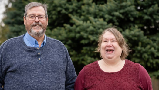 Un homme et une femme se tiennent côte à côte à l'extérieur, devant un grand conifère ; tous deux sourient et rient. L'homme, à gauche, a les cheveux gris, une barbe et porte des lunettes ; il est vêtu d'un pull Henley bleu marine par-dessus une chemise bleu clair. La femme, à droite, a les cheveux châtain clair et porte un haut texturé bordeaux foncé. L'image se présente sous la forme d'une grande bannière.