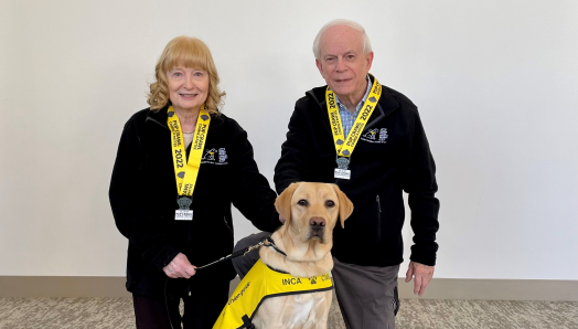 Mary et John Crocker, tous deux souriants, sont agenouillés par terre à côté d'un chien blond portant un gilet du CNIB portant l'inscription « Futur chien-guide ».