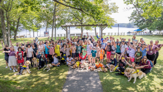 Au Camp Lake Joe, un large groupe de personnes de tous âges, dont bon nombre sont accompagnées de leurs chiens compagnons ou tiennent une canne blanche, lèvent joyeusement les mains en l'air, le lac en toile de fond.
