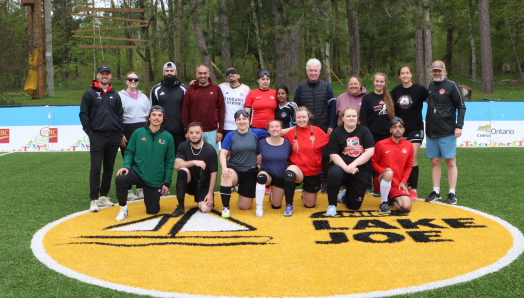 Un groupe de joueurs de soccer aveugles, de tailles, d'âges et de couleurs de peau variés en tenue de sport se tient derrière un ballon de cécifoot sur le terrain de soccer accessible du camp Lake Joe d’INCA.