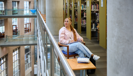 Ashlyn, vêtue d'un pull rose et d'un jean, est assise sur une chaise en bois près de la balustrade en verre d'une bibliothèque à plusieurs étages. Elle tient un cahier et un stylo sur ses genoux et regarde par-dessus le bord de la balustrade.