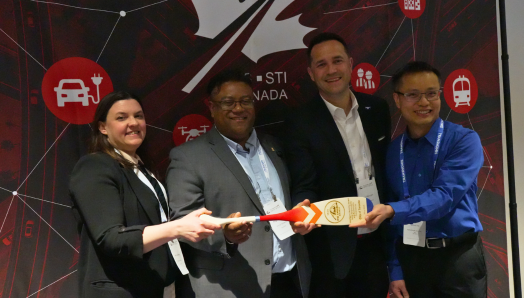 Quatre membres de l'équipe du projet pilote « Zones de construction de trottoirs accessibles aux personnes aveugles et à basse vision » posent ensemble lors d'un événement, souriants et tenant leur prix, une pagaie en bois sur laquelle sont gravées les informations relatives à cette distinction. Mary Ann, à l'extrême gauche, tient une extrémité de la pagaie tandis que les autres membres de l'équipe se rassemblent autour d'elle.