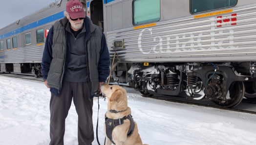 Dan se tient dans la neige aux côtés de son chien-guide, Sparky, un labrador blond, qui est assis, son harnais aux épaules, et lève les yeux vers lui. Un train de voyageurs VIA Rail est à l’arrêt sur les voies derrière eux.