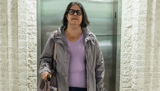 Karoline se tient à côté de son chien-guide, un labrador noir nommé Guardian, devant un ascenseur. Guardian porte son harnais ; Karoline tient la poignée du harnais dans sa main gauche et la poignée d’une valise dans sa main droite.