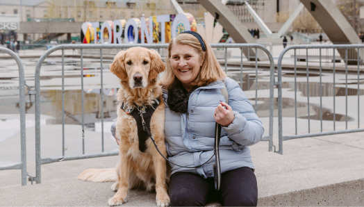  Lindsay est assise à côté de son chien-guide, Charles, un golden retriever, sur la place Nathan Phillips à Toronto. Charles porte son harnais et Lindsay sourit à la caméra, un bras passé autour de lui. 