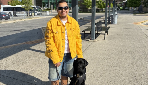 Tye se tient aux côtés de Zed, son chien-guide labrador noir, dans une station de transport en commun en plein air. Tye tient le harnais et la laisse de Zed.
