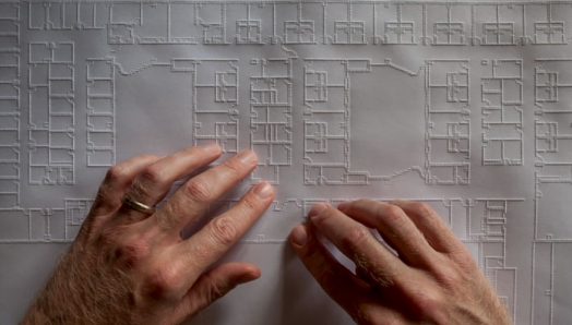 Deux mains parcourent un grand plan architectural tactile. Des lignes en relief délimitent les pièces, les couloirs, les murs et d'autres éléments de la disposition du bâtiment.