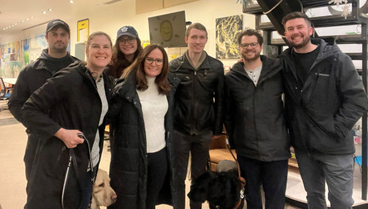 Une photo de groupe de Larissa et Shannon en compagnie de quatre membres de l'équipe de production de la vidéo de la Police de l'Ontario et de Jack, un utilisateur d'un chien-guide qui apparaît dans la vidéo, au GTA Community Hub. Larissa et Jack sont accompagnés de leurs chiens-guides.